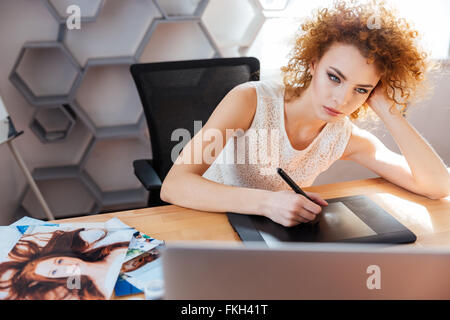 Concentrated pretty redhead young woman photographer processing pictures using graphic tablet in office Stock Photo