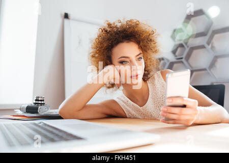 Relaxed cute curly young woman leaning on the table and using mobile phone in the office Stock Photo