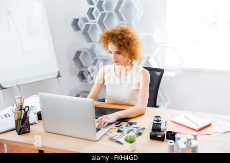 Beautiful curly young woman photographer sitting on workplace and using laptop with graphic tablet Stock Photo