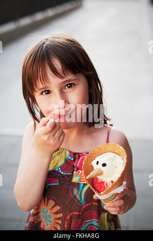 Cute girl eating ice-cream  outside in hot summer day. An ice-cream is made like a snowman with eyes and nose. Stock Photo