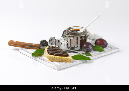 jar of plum jam, fresh and dried plums and slice of baguette on wooden cutting board Stock Photo