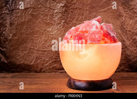 Turned on Himalayan pink salt lamp carved as a bowl on a wooden table with wrinkled parchment paper background. Stock Photo