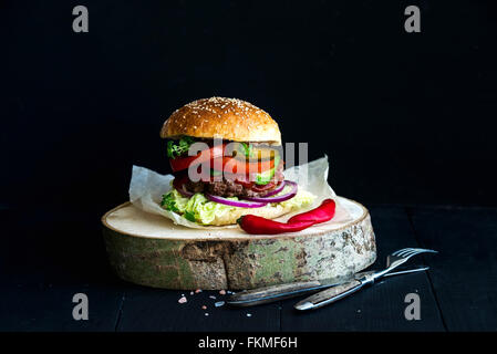 Fresh homemade burger on wooden serving board with red chili peppers over black  background Stock Photo