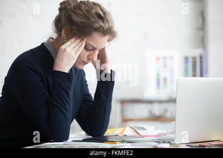 Portrait of young stressed woman sitting at home office desk in front of laptop, touching head with frustrated facial expression