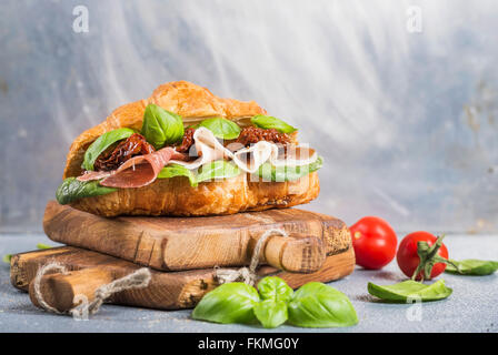 Croissant sandwich with smoked meat Prosciutto di Parma, sun dried tomatoes, fresh spinach and basil on stone textured grey back Stock Photo