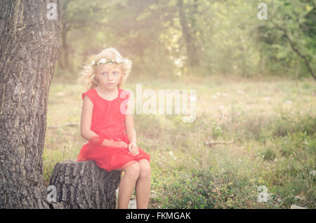 little blond girl in red dress sitting in the tree trunk Stock Photo