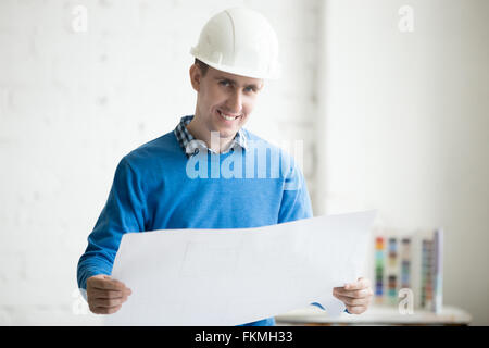 Portrait of young cheerful handsome engineer in white helmet holding blueprint, standing in architectural agency office Stock Photo