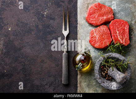 Raw fresh marbled meat Steaks with seasonings and meat fork on stone slate on dark background Stock Photo