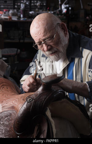 An old saddle craftsman works by window light in his workshop. Stock Photo