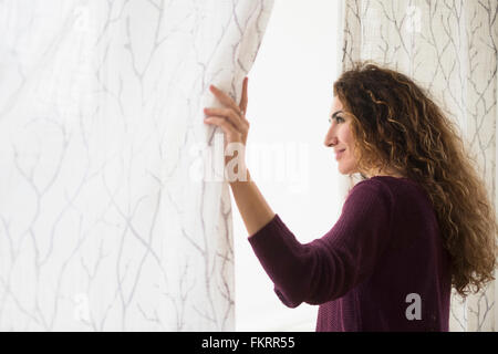 Mixed race woman looking out window curtain Stock Photo