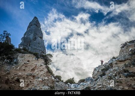 Low angle view   young man standing on cliff pointing at cone shape rock formation, Ogliastra, Sardinia, Italy Stock Photo