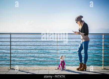 Full length side view   young woman with dog by railings in front   ocean using smartphone Stock Photo