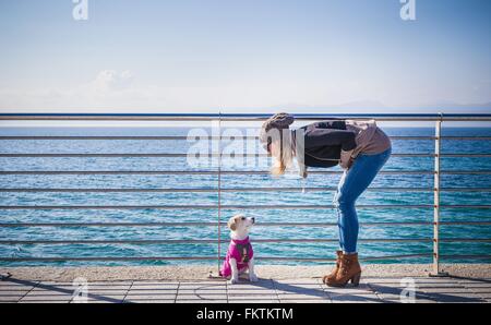 Full length side view   young woman by railings in front   ocean bending over looking at dog Stock Photo