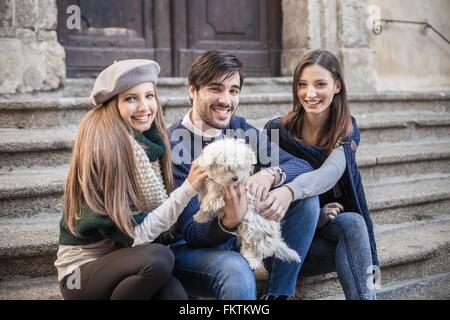Friends sitting side by side on steps holding fluffy dog looking at camera smiling Stock Photo