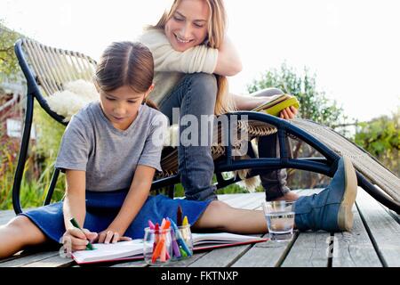 Girl drawing on decking with mother sitting close by, smiling Stock Photo