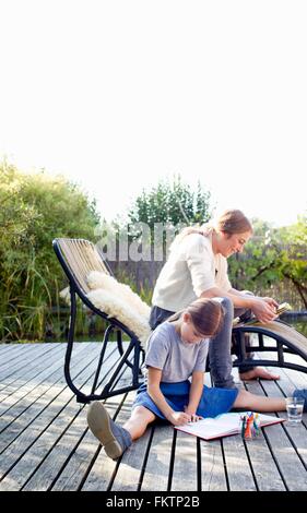 Girl drawing on decking with mother sitting on lounge chair Stock Photo