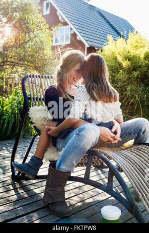 Daughter sitting on mother's lap on lounge chair on wooden decking Stock Photo