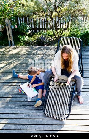 Mother using tablet, daughter drawing on wooden decking Stock Photo