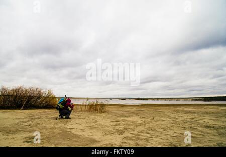 Young man hiking, crouching, taking photograph of lake view, rear view Stock Photo