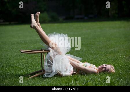 Portrait of young woman lying on grass, sitting on fallen stool, smiling Stock Photo