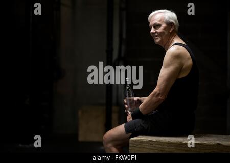 Senior man sitting with water bottle in dark gym Stock Photo