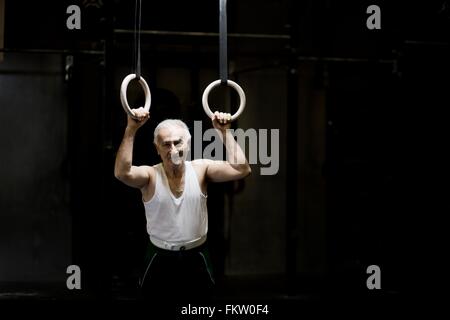 Portrait of grey haired senior man holding gym rings in dark gym