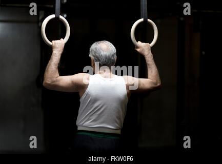 Rear view of senior man holding gym rings in dark gym