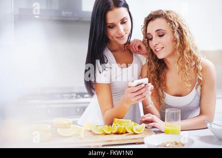 Two young women reading smartphone text at kitchen counter Stock Photo
