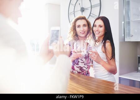 Young man photographing female friends on smartphone in kitchen Stock Photo