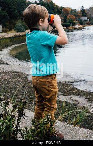 Young boy standing beside lake, looking through binoculars Stock Photo