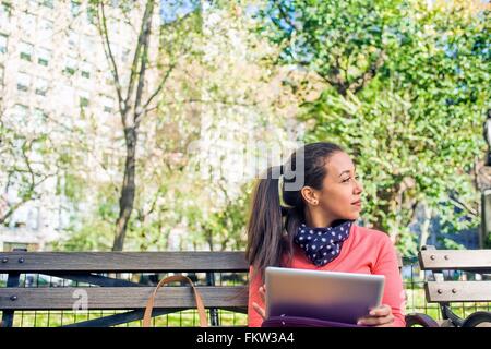 Young woman looking sideways from park bench whilst using digital tablet Stock Photo