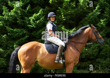 Side view of mature woman on horseback wearing riding hat and boots looking at camera smiling Stock Photo