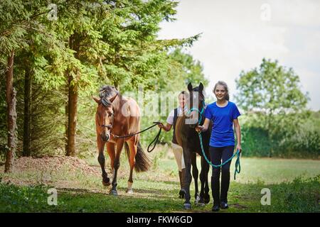 Full length front view of girl and mature woman holding lead rope, walking with horses looking at camera smiling Stock Photo