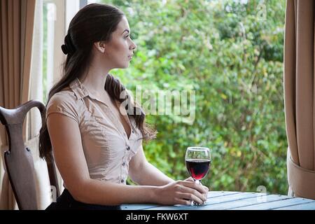 Young woman sitting at table in front   window holding glass   wine looking away Stock Photo