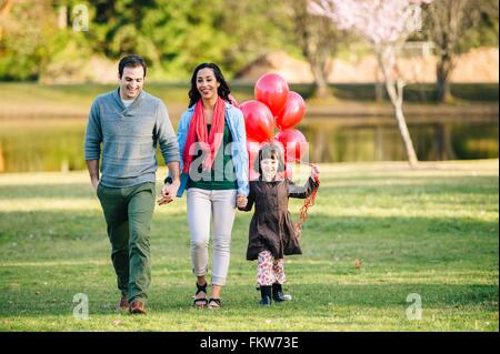 Young couple and daughter with bunch of red balloons strolling in park Stock Photo
