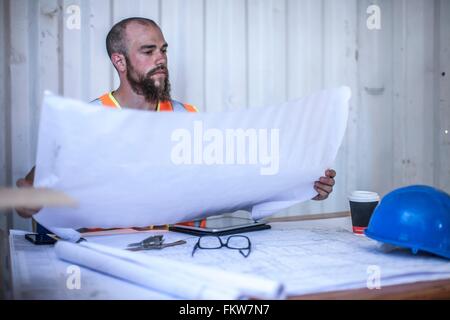 Construction worker sitting at desk looking at blueprint in portable cabin Stock Photo