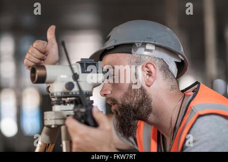 Young male surveyor looking through theodolite giving thumbs up on construction site Stock Photo