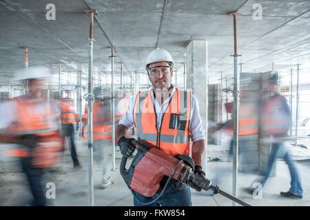 Mid adult male builder holding building equipment  on busy construction site Stock Photo