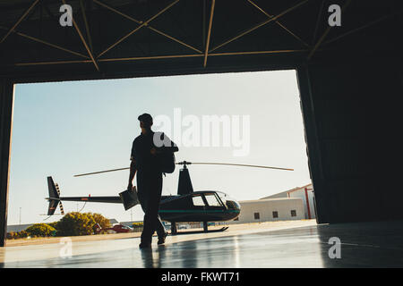Silhouette of a pilot arriving at the airport with a helicopter in background. Helicopter pilot in airplane hangar. Stock Photo