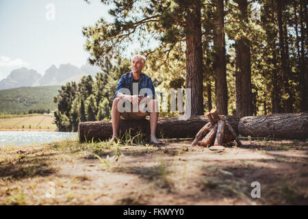 Portrait of a mature man sitting on a log staring out to the lake. Senior man camping by the lake on a summer day.
