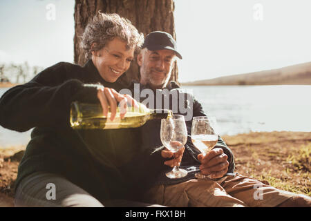 Senior couple enjoying drinks at campsite near Lake. Mature woman pouring wine in glasses, both sitting under a tree on a summer Stock Photo