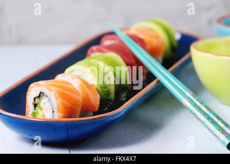 Sushi set salmon and avocado rolls served in bright ceramic plates with chopsticks and soy sauce on blue wooden table. Stock Photo