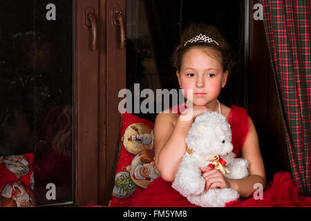 Thoughtful adorable little girl, dressed in a lush red gown, hugging a plush bear next to the window Stock Photo