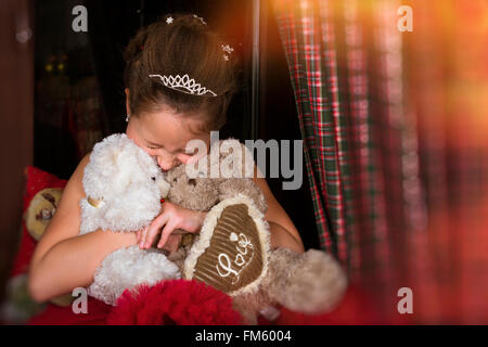 Smiling adorable little girl, dressed in a lush red dress, much hugging two plush bears, heart with inscription Love Stock Photo