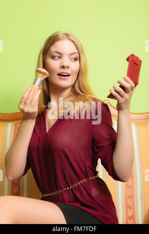 Young attractive woman applying make up with powder brush. Pretty gorgeous girl sitting on vintage retro sofa couch. Fashion. Stock Photo