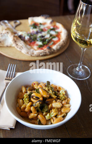 A bowl of white bean, romanesco and mussels soup with white wine and a pizza on the table behind it.