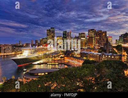 bright lights of SYdney city CBD at sunset as viewed from Harbour Bridge pylon towards Circular quay with docked passenger ship Stock Photo