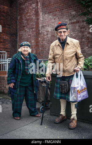 A senior man and woman in traditional Scottish dress pause for a breath on the street in front of a brick wall. The woman has a Stock Photo
