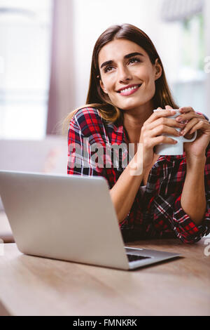 Smiling thoughtful woman holding coffee cup with laptop Stock Photo