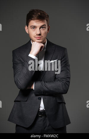 Serious attractive young businessman in black suit standing with hands folded over grey background Stock Photo